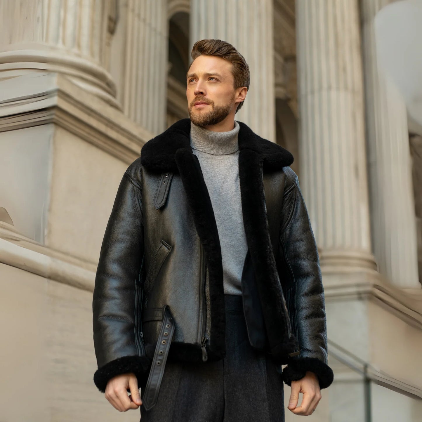 Man wearing a black leather jacket with fur trim standing in front of classical architecture.